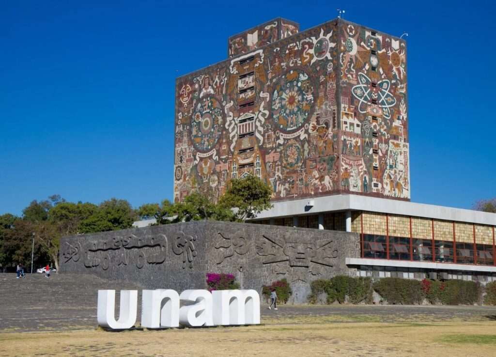 Three quarter view of the UNAM Central Library from the main lawn featuring large white "UNAM" letters in the foreground, a relief carved volcanic rock terrace wall, and the mosaic tower under a clear blue sky.