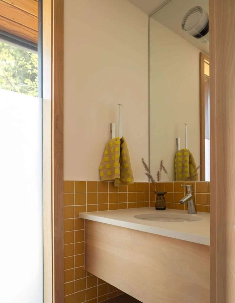 Modern bathroom vanity with warm mustard yellow tiles, light wood cabinets, and a clean white countertop.