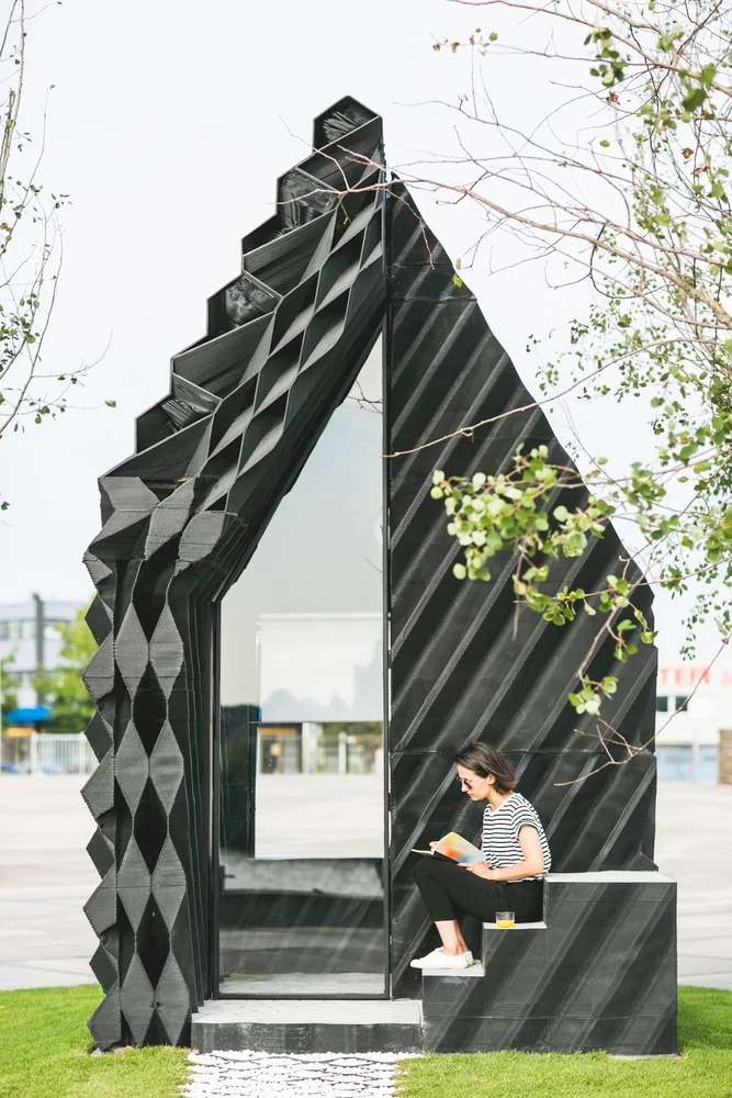 A woman sitting on concrete porch steps of a black 3D-printed tiny cabin while reading a book.