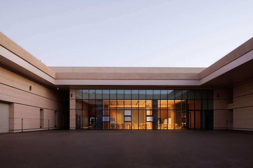 Ground-level view of a sunken courtyard plaza featuring a large glass curtain wall facade under a clear evening sky.