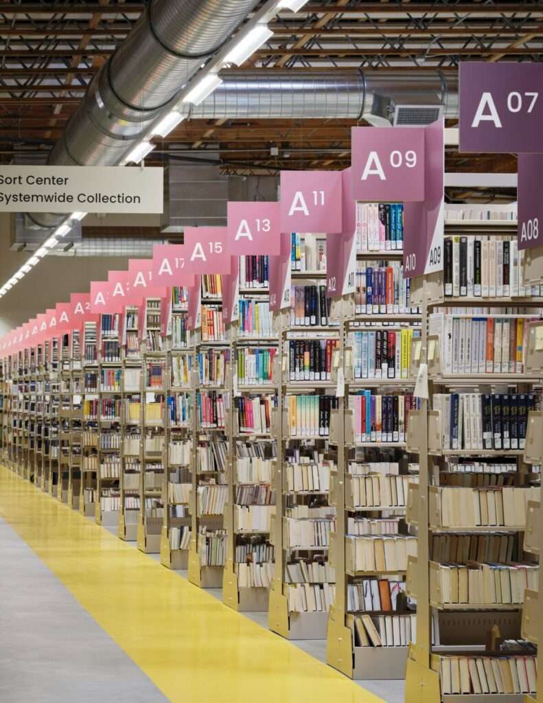 Looking down a long aisle of library book stacks with pink and purple row markers under industrial light fixtures and large exposed overhead ventilation ducts.