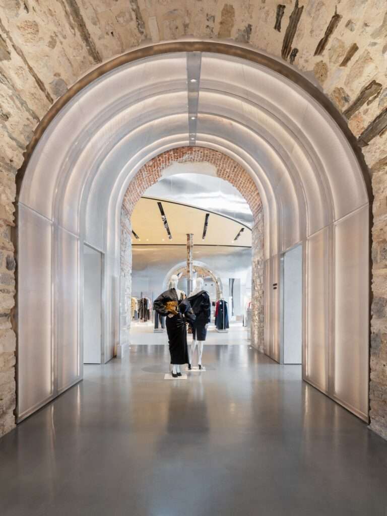A symmetrical view through a stone archway fitted with a backlit curved mesh lining, framing fashion mannequins inside the store.