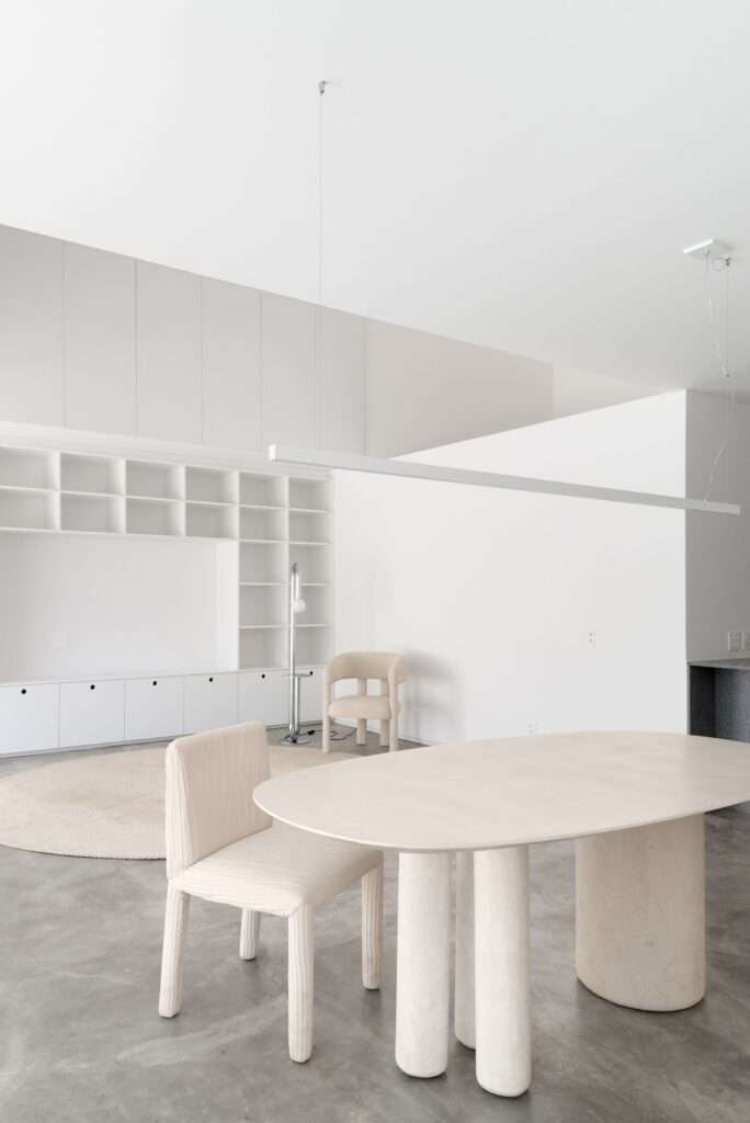 Bright white minimalist interior dining area with a modern stone oval table, textured chairs, a built-in white shelving unit, and a linear pendant light over a polished concrete floor.