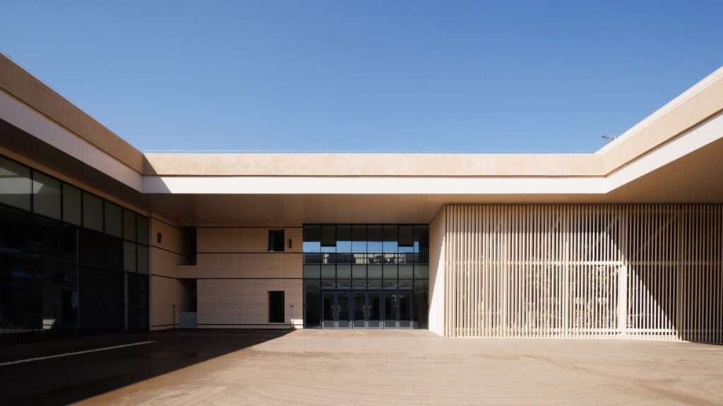 Courtyard area within a modern building showing vertical wooden slats flanking a recessed glass entrance under a clear blue sky.