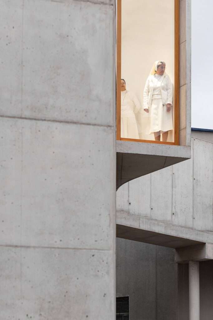 Close-up of a cantilevered concrete volume with a large wood-framed window, showing two women in white garments looking out from the interior.