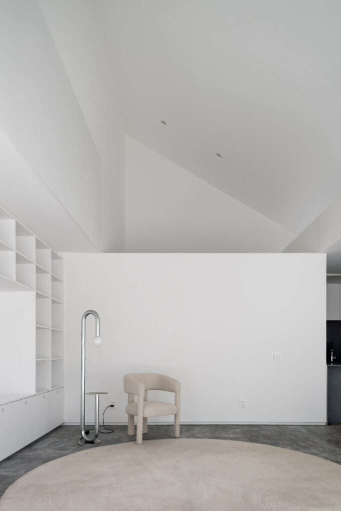 Minimalist interior room with high sloped ceilings reflecting the butterfly roof form, a single beige accent armchair, a circular rug, and a modern tubular chrome floor lamp.