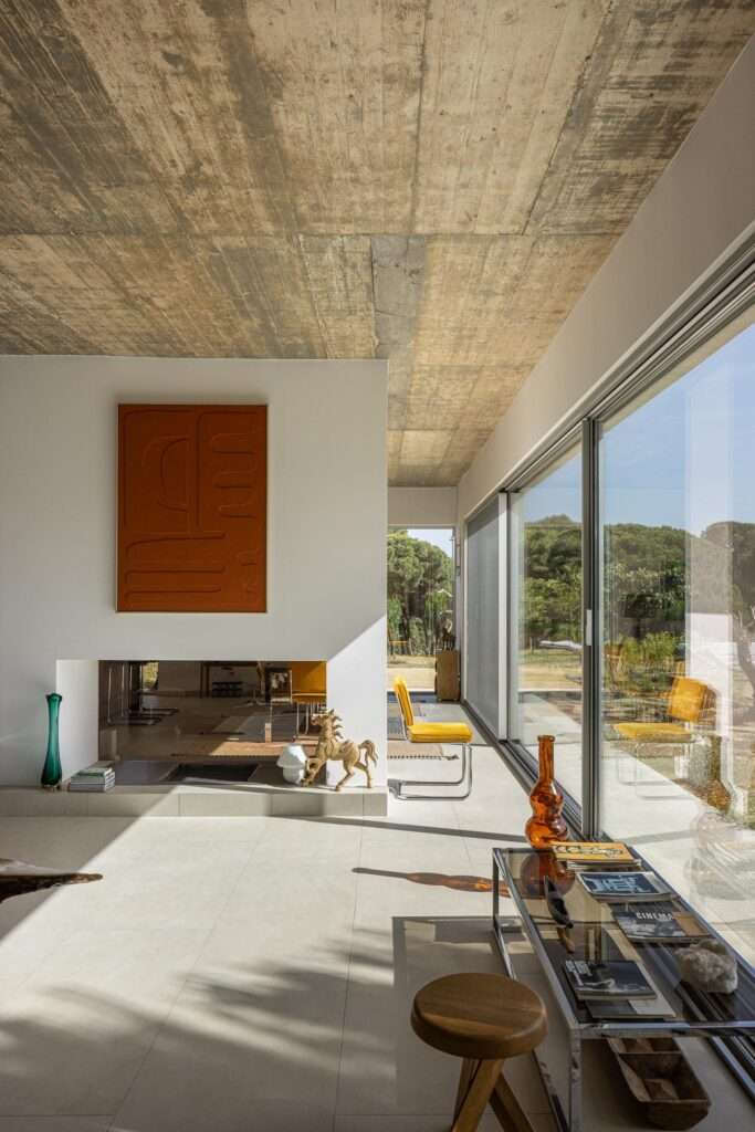 Long minimalist interior hallway with an exposed concrete ceiling, glass windows on the left showing an internal courtyard, and white walls leading to a black lounge chair in the distance.