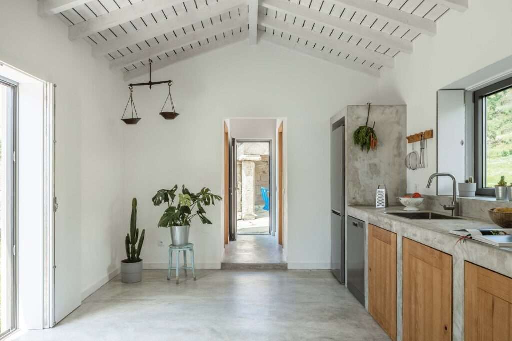 Minimalist kitchen and corridor path within Abadim House showcasing polished concrete floors, raw concrete structural elements, and a view through a doorway to an old stone exterior wall.