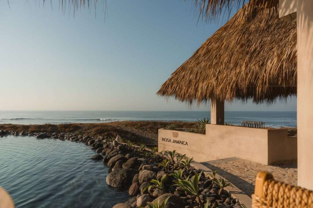 The concrete entrance wall of Rosa Jamaica Beach Club, integrated with a volcanic stone retaining barrier next to an organic water pool and a thatched pavilion.