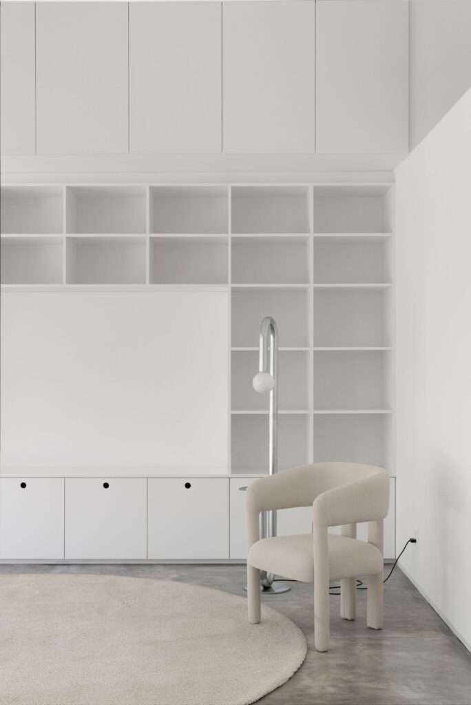 Close-up of custom white modular bookshelves and cabinets with a contemporary beige armchair and a curved chrome floor lamp on a gray concrete floor.