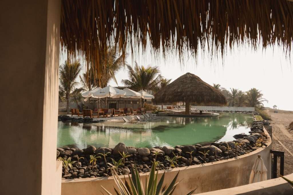 A wide scenic view of a curved swimming pool surrounded by sand, white umbrellas, wooden lounge chairs, and palm trees under a bright sky.