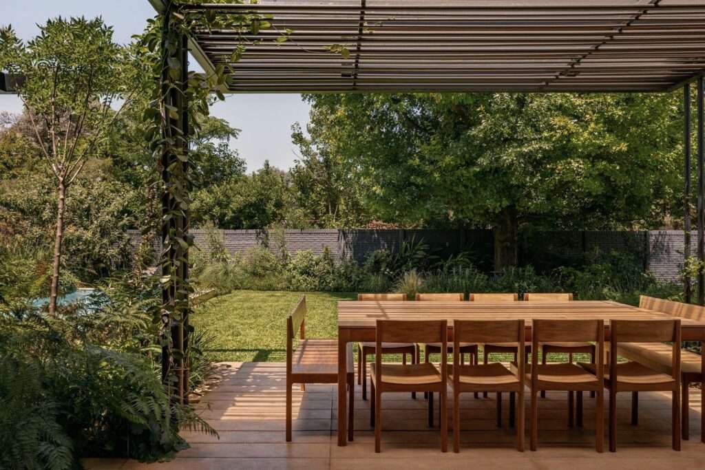 Covered outdoor dining area with a wooden table, surrounded by lush garden greenery and a view of the pool.