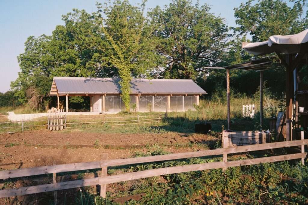 Wide shot of the Gardeners' Shelter in a rural landscape with wooden fences and cultivated agricultural plots in the foreground.