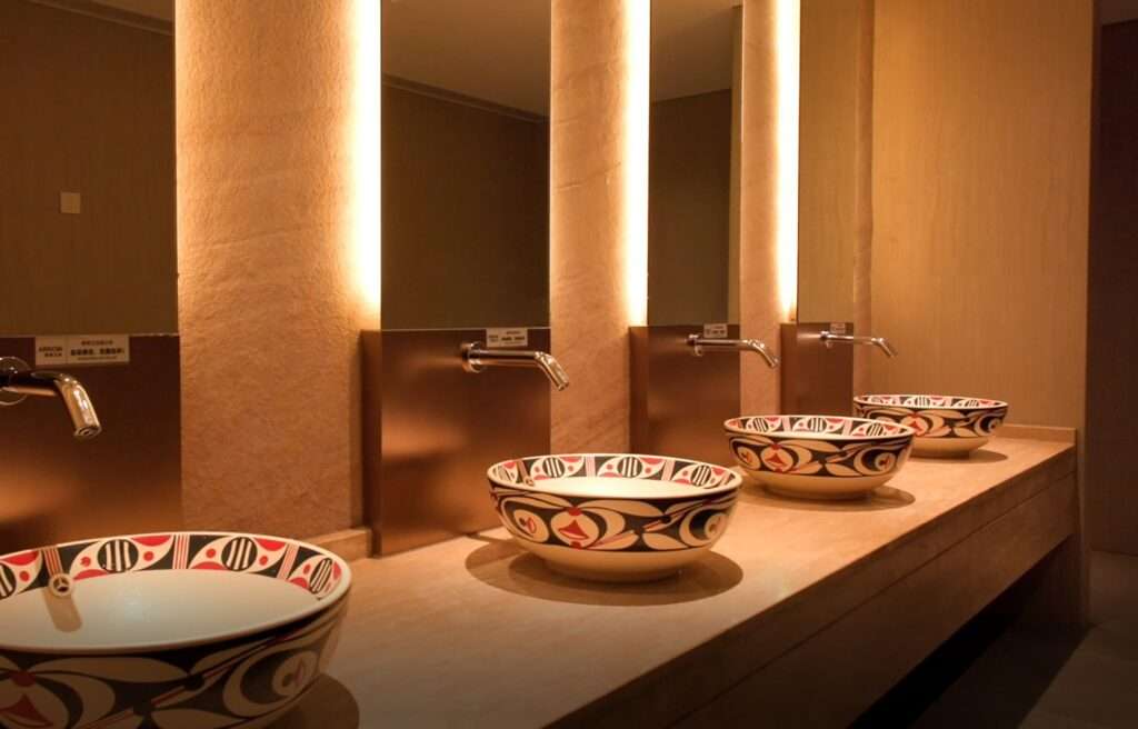 Indoor view of a contemporary public restroom with decorative ceramic washbasins, metallic plumbing fixtures, and backlit mirrors against stone pillars.