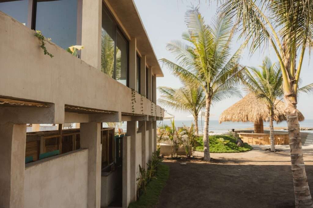 A two-story brutalist-inspired raw concrete building with wide windows, overlooking an outdoor courtyard with palm trees and a thatched bar hut.