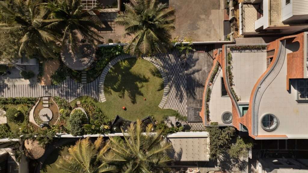 Full frontal elevation of a multi-story red brick residential building featuring organic, flowing curves, terraced balconies, and a landscaped front lawn with a walkway in Ahilyanagar.