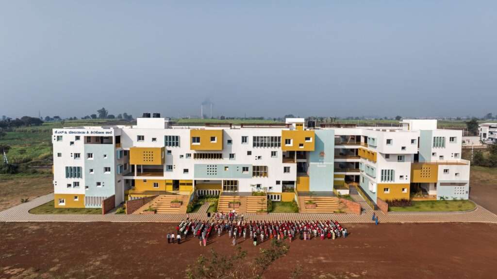 High-angle drone overview of a large decentralized school building showing the asymmetric layout, internal courtyards, and a large group of students assembled on the ground field.