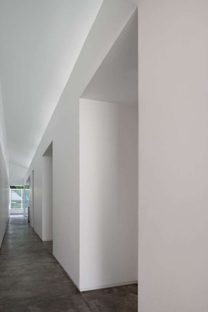 Long, minimalist white interior hallway with polished concrete floors and geometric door openings leading toward an outdoor courtyard view at the far end.