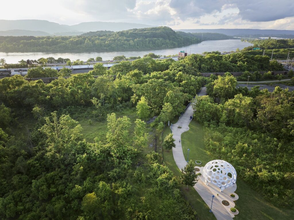 High-angle aerial drone shot of the Moonrise pavilion situated alongside a river pathway surrounded by lush green forests and hills.