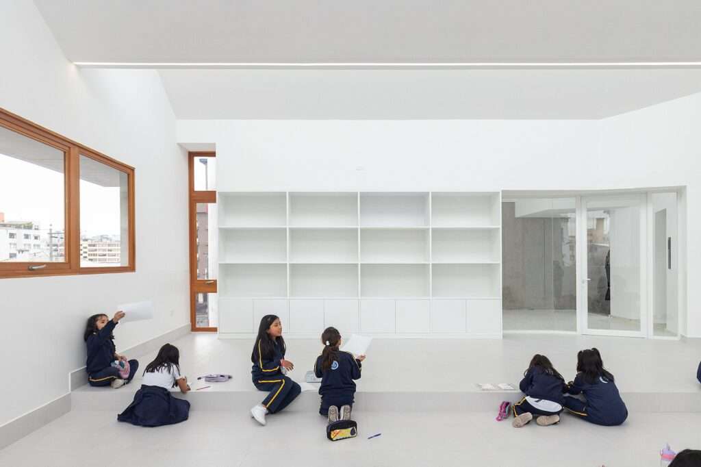 Bright white interior art studio with children sitting on a raised platform floor, drawing, next to a large empty white bookshelf unit.