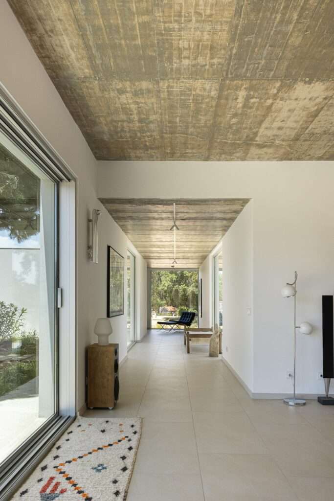 Exterior evening view of the interconnected white geometric volumes of Melides House, showing illuminated minimalist interiors through large windows amidst wild grass.