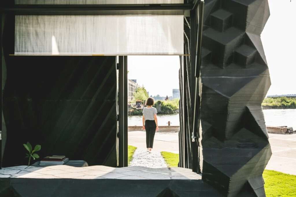 Looking through the window of a dark 3D-printed micro cabin at a woman walking out toward an open river waterfront.