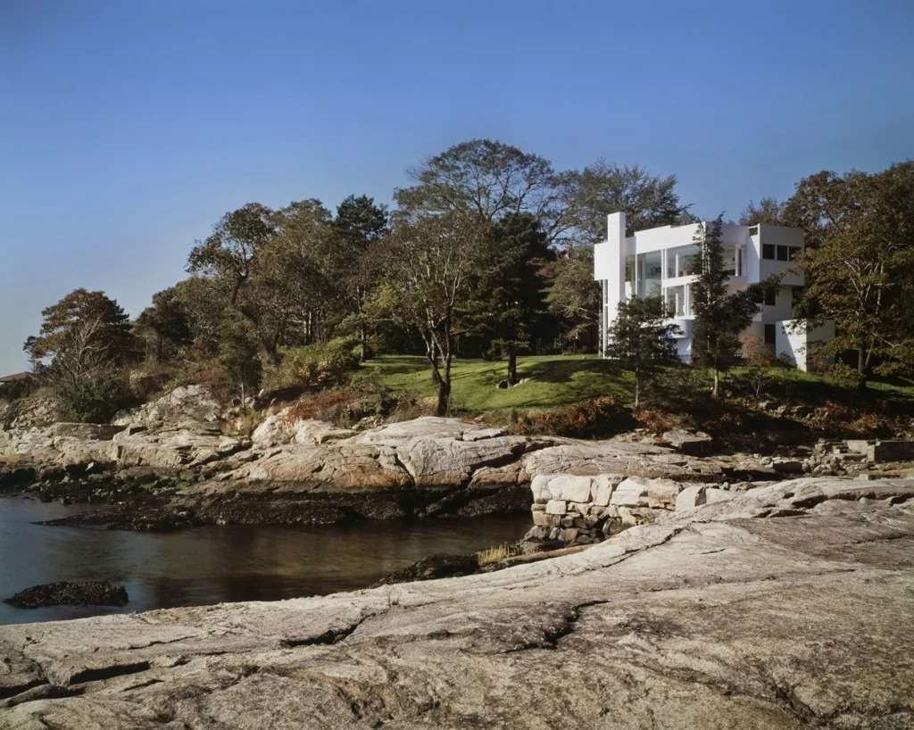 Wide shot of the white modernist Smith House standing on a rocky coastal shoreline surrounded by dense trees.
