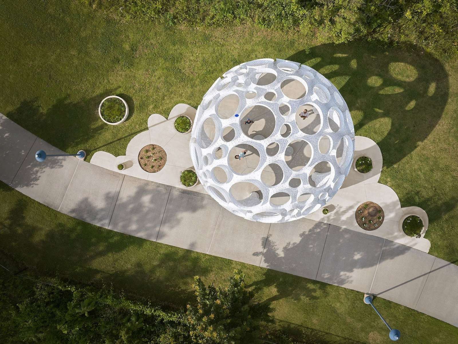 Direct overhead aerial view of the spherical Moonrise pavilion, its circular landscape footprint, concrete walkways, and cast shadows on the grass.