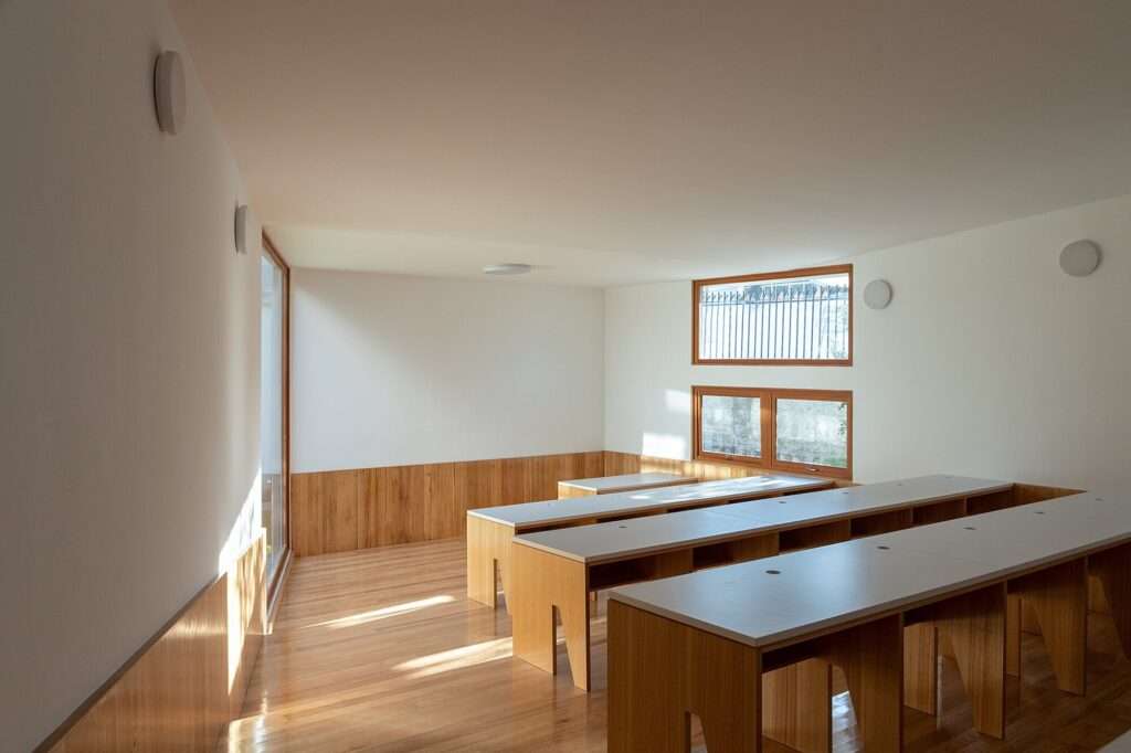 Interior classroom view featuring warm wood flooring, wood wall paneling, and built-in wooden desks under soft natural light from square windows.