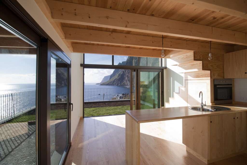 An exterior view from an upper perspective looking down at the gabled wooden house at Jardim do Mar during sunset. The house is built on a stone-walled slope, with a pedestrian path and stairs leading towards the rocky ocean coastline in the background.