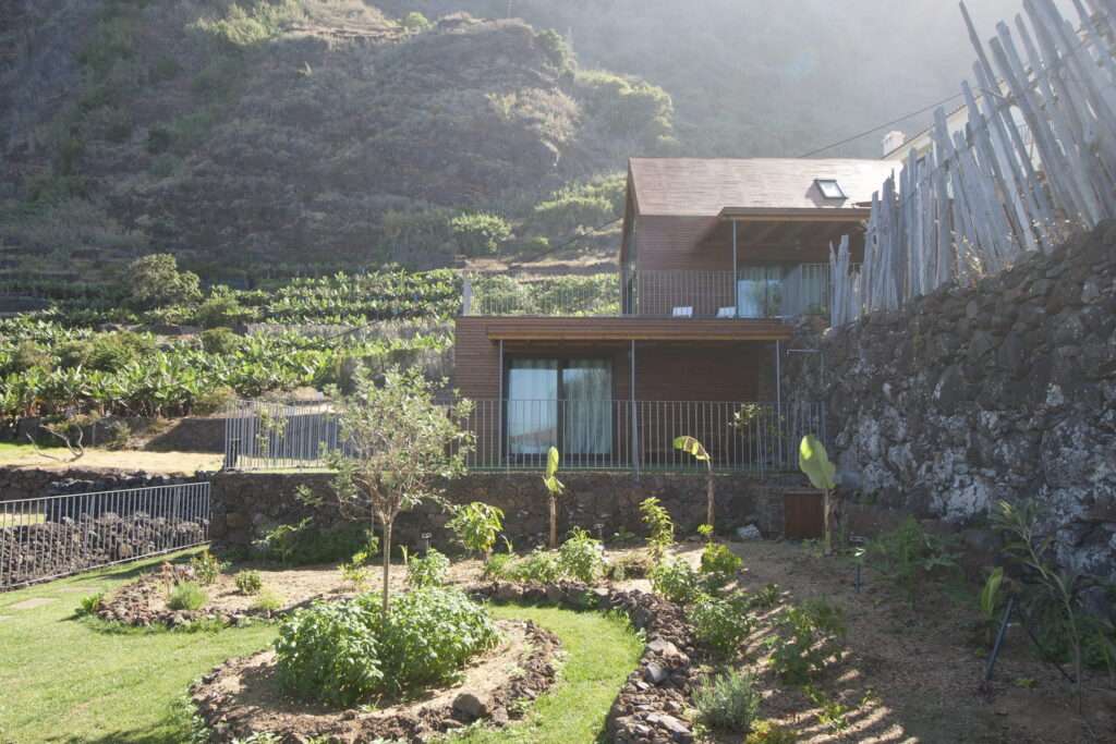 An interior photograph of the lower apartment in the Jardim do Mar houses, viewed from the living area through large sliding glass doors. The interior features light wood floors, built-in wooden kitchen cabinetry, and an open plan. The doors open to a grassy terrace with a view of a steep coastal cliff and the ocean.