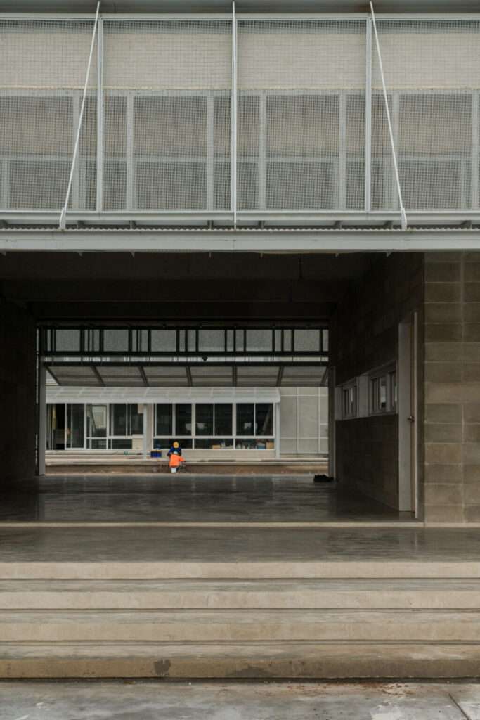 Open-air breezeway of the LPP Rice Grading Center showing raw concrete steps, structural columns, and an overhead steel mesh canopy optimizing natural ventilation.