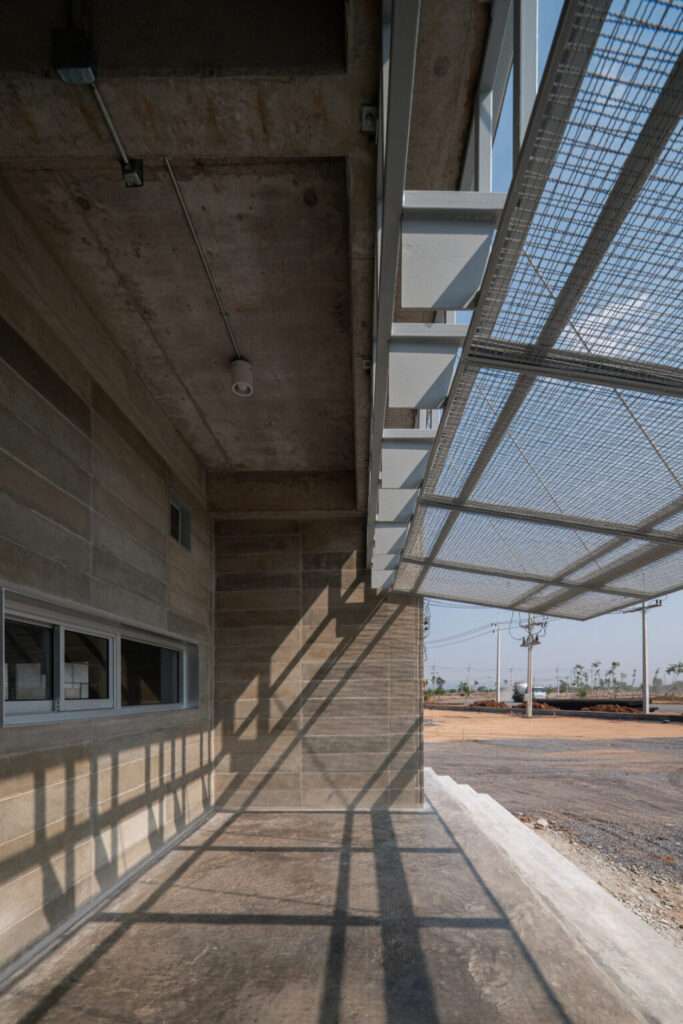 Profile view of an industrial building facade featuring custom concrete blocks made from rice husks, a horizontal window, and a steel mesh awning casting shadows.
