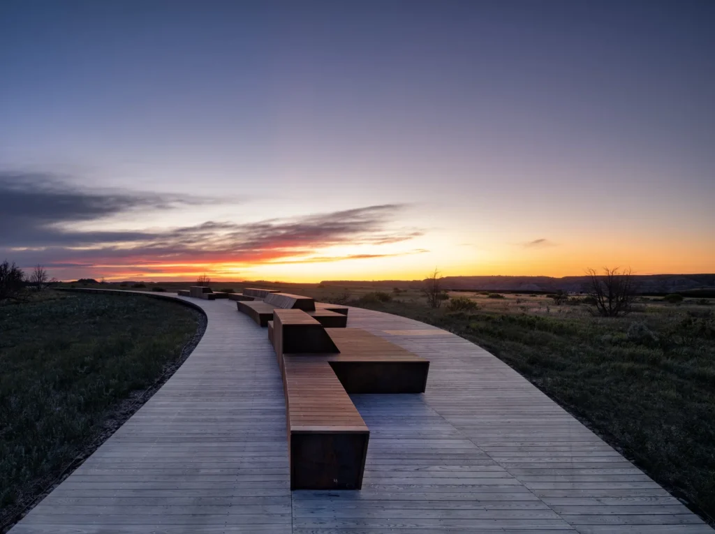 An elevated wooden boardwalk with integrated seating modules curves through a prairie landscape at sunset.
