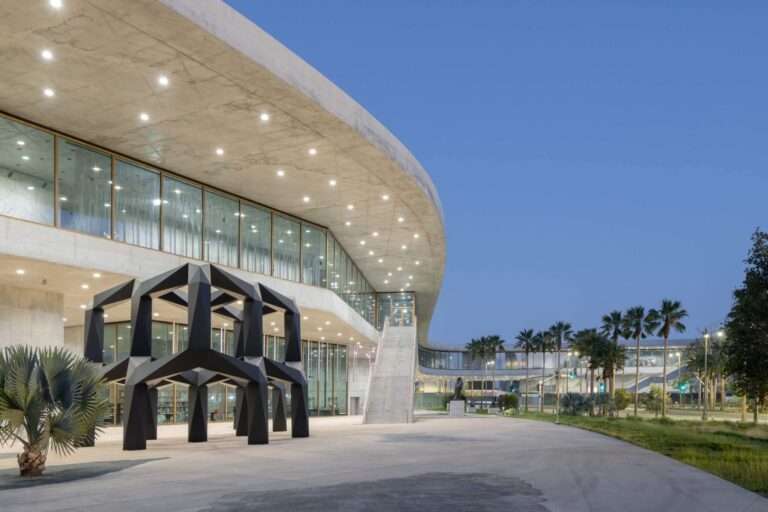 A low-angle exterior nighttime view of a concrete canopy and glazed facade overlooking an outdoor plaza with a black geometric sculpture.