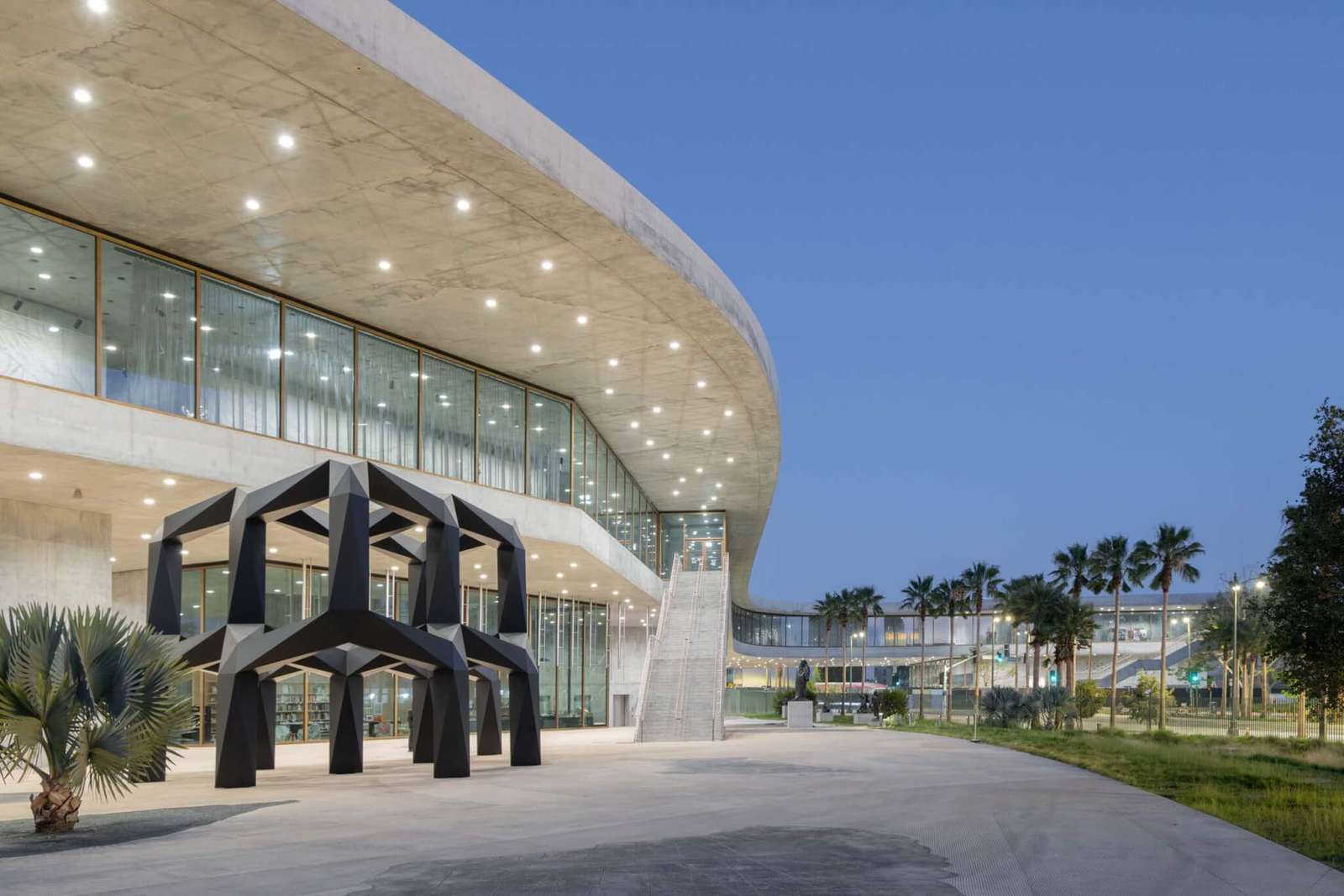 A low-angle exterior nighttime view of a concrete canopy and glazed facade overlooking an outdoor plaza with a black geometric sculpture.