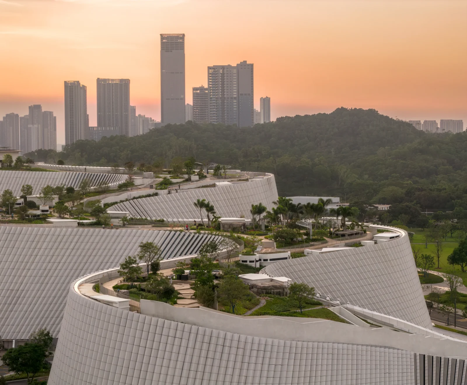 Stepped green rooftop terraces with winding walkways on a curving building facade overlooking city skyscrapers.