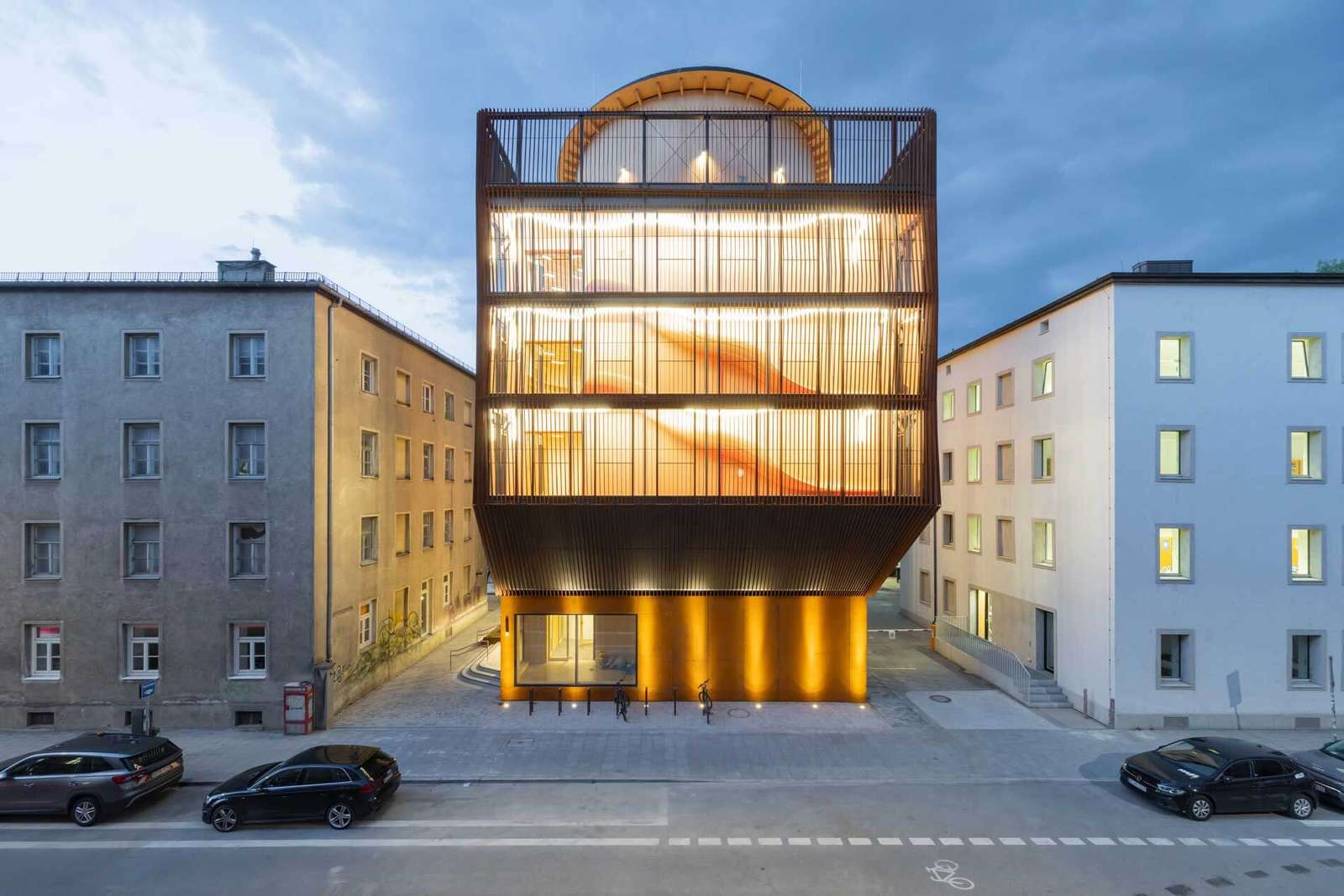 Symmetrical street view of an illuminated modern five-story timber kindergarten wedged between two urban residential buildings at dusk.