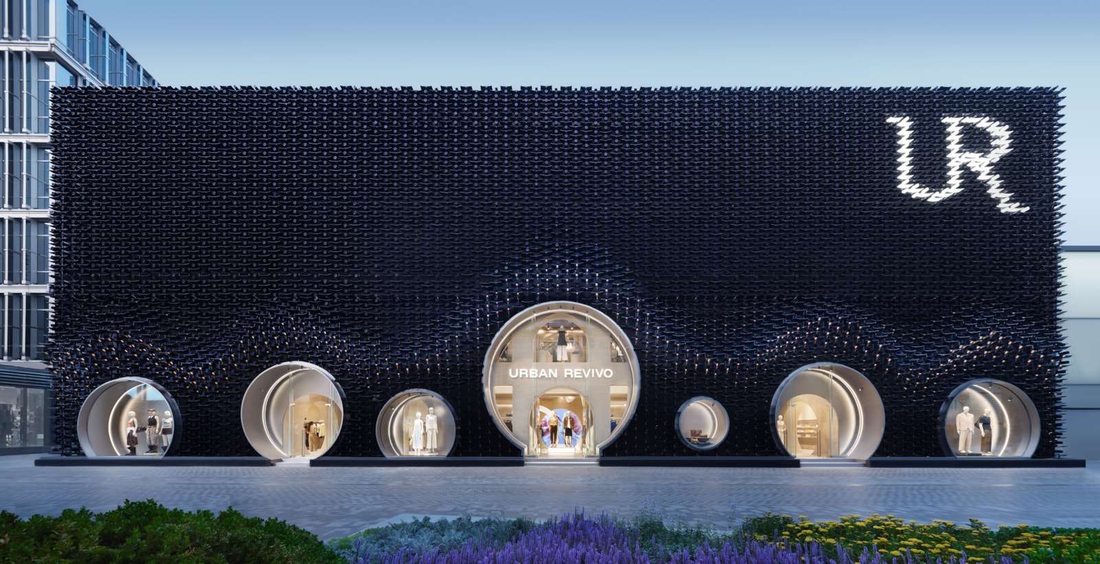 A wide nighttime shot of a dark textured facade with circular window displays and illuminated signage at the top right.