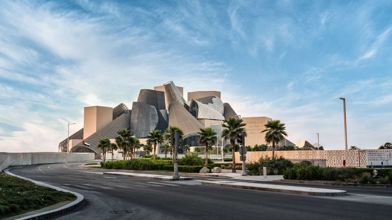 An asphalt road curving toward the complex architectural massing of Guggenheim Abu Dhabi under a blue sky.
