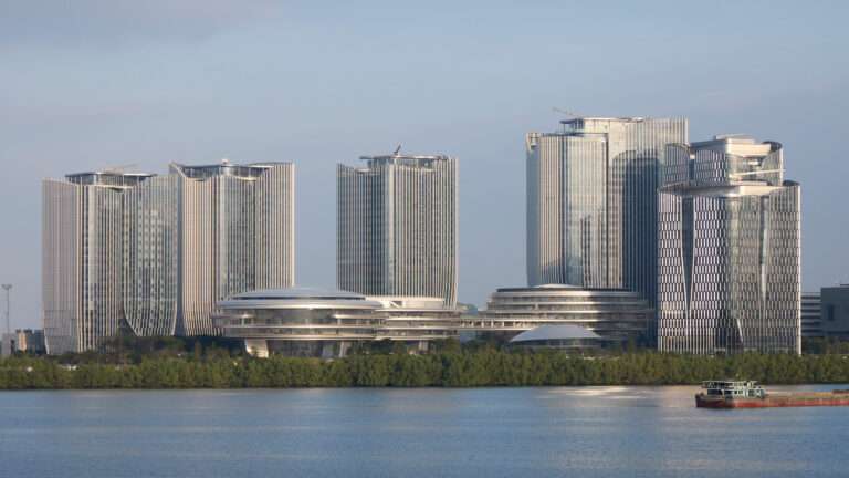 An aerial view of an architectural complex with three rounded cloud-like buildings and several skyscrapers at sunset.