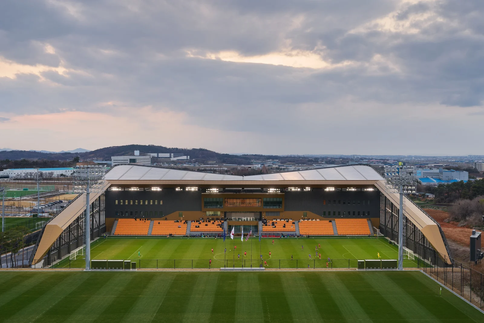An elevated view of an outdoor football pitch and main stand with yellow seating under a cloudy sky.