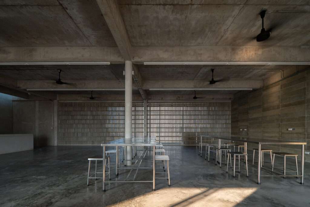 Interior of the LPP communal canteen with exposed raw concrete ceilings, industrial fans, stainless steel dining tables, and a perforated brick screen wall allowing natural light.