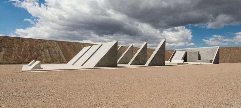 A series of sharp, triangular concrete monolithic structures arranged within a vast gravel desert plateau under cloudy skies.