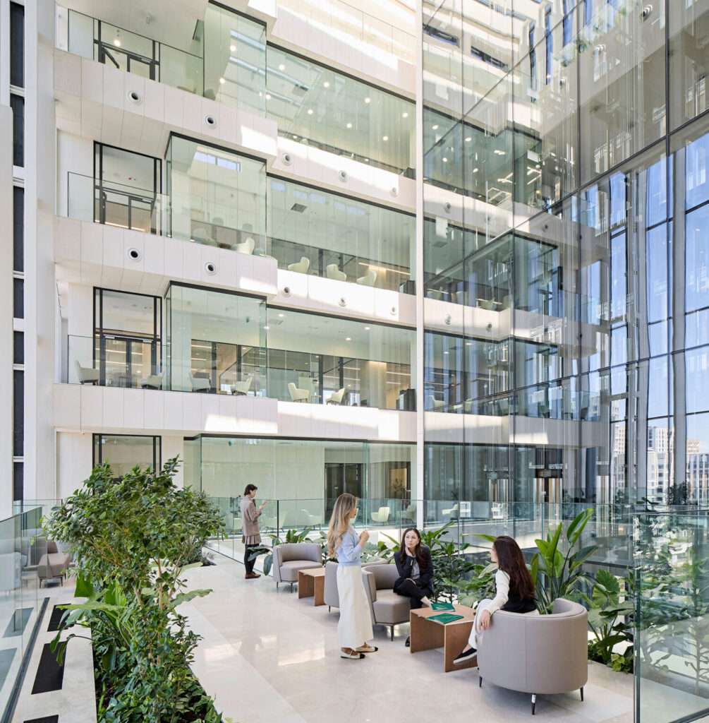 A multi-story glazed atrium interior with multi-level office terraces, indoor plants, and people seated.