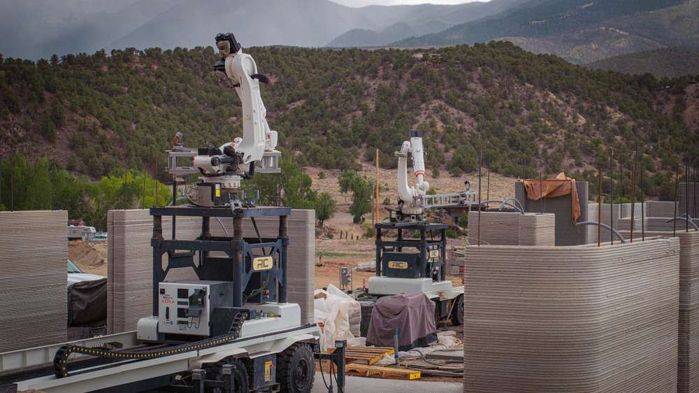 Two autonomous robotic printing arms on mobile bases positioned next to layered concrete walls.