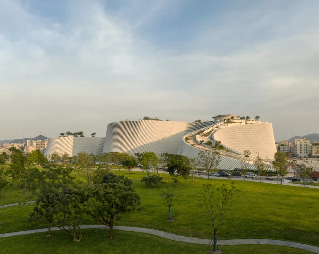 Curved building with sloped granite-clad facade modules rising from a green park field.