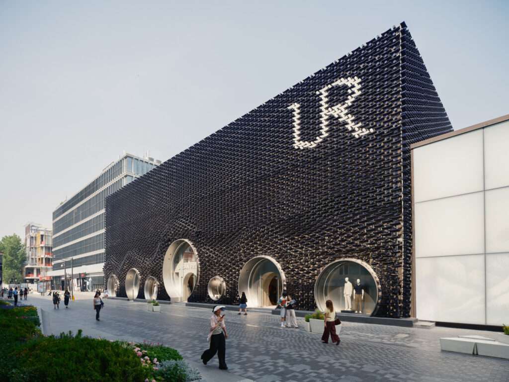 Daytime perspective view of a long dark facade with circular portals along a pedestrian street.