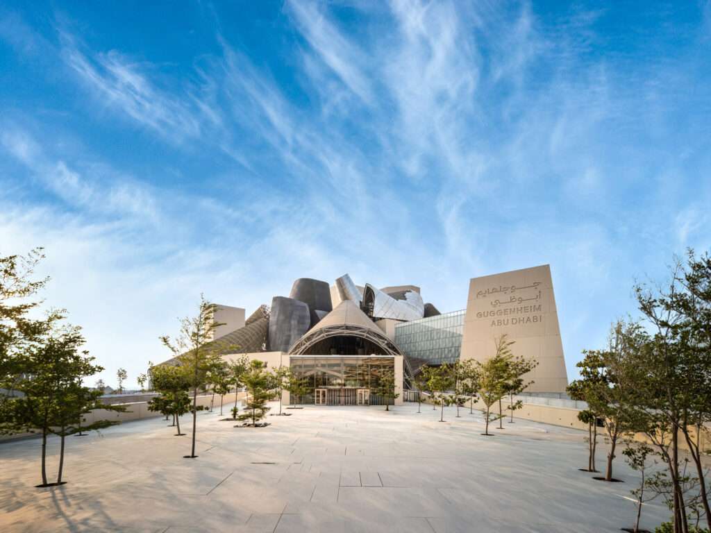 A wide paved plaza lined with young trees leading to the glazed entrance hall of Guggenheim Abu Dhabi.