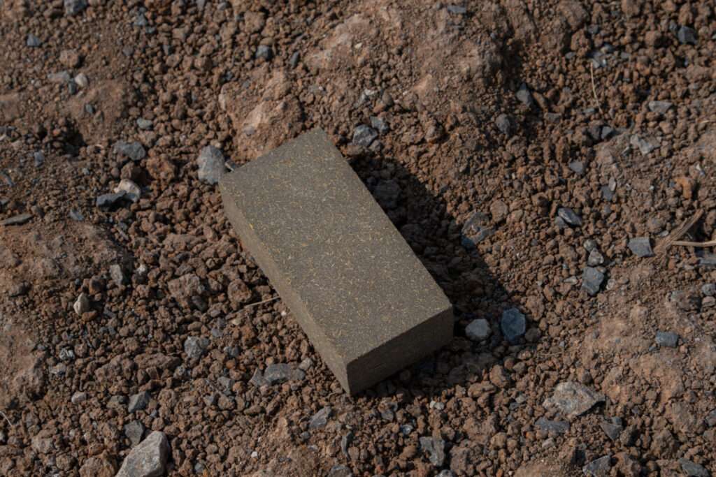 Close-up of a single sustainable bio-concrete rectangular block containing embedded rice husks and agricultural waste, sitting on dark soil.